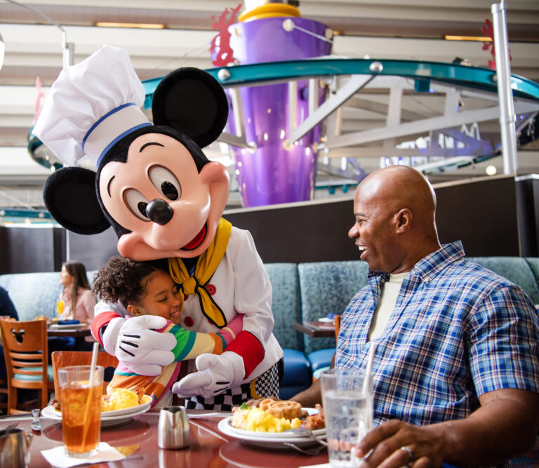 Mickey Mouse hugging child at Chef Mickeys with Dad smiling looking on